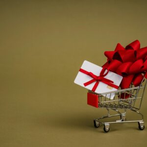 Mini shopping cart containing a gift card adorned with a red bow on a neutral background, ideal for retail concepts.